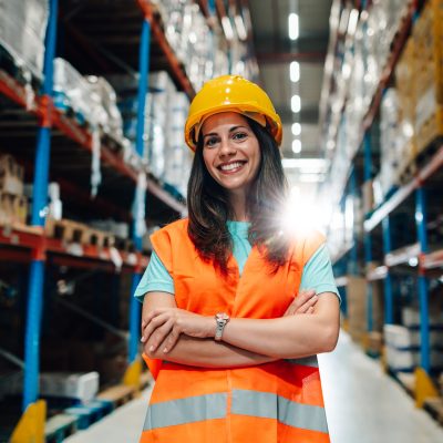 Female supervisor wearing a safety vest and hard hat smiles while standing with crossed arms in a warehouse aisle, demonstrating confidence and professionalism in her role