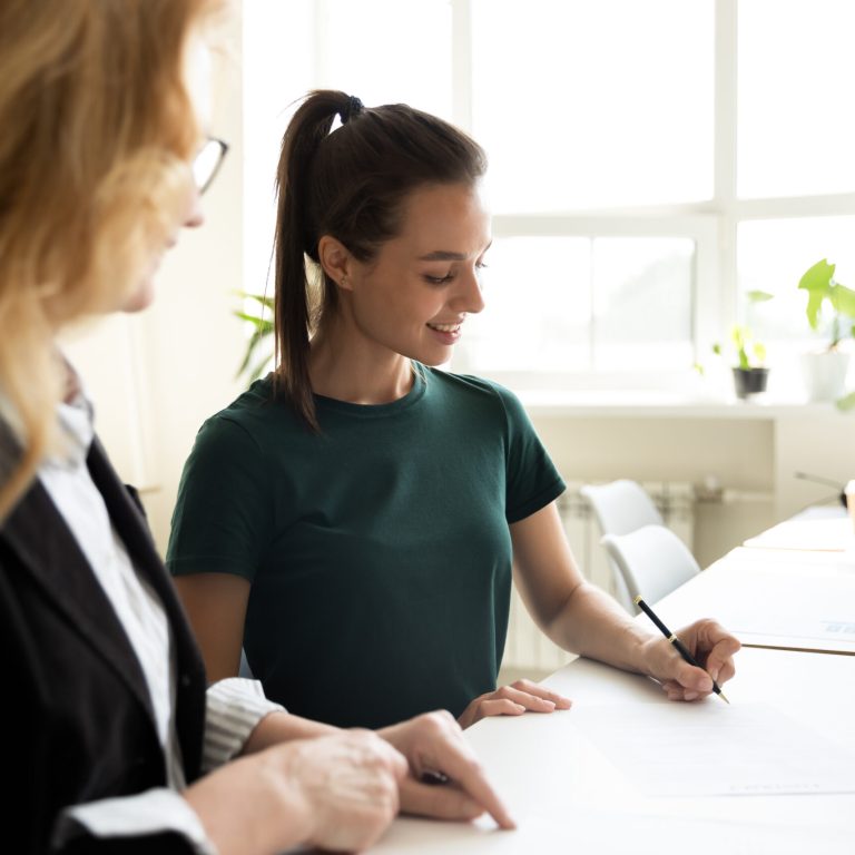 Mature business teacher supervising happy student writing notes. Middle aged mentor training intern at office workplace. HR manager helping new employee to fill and sign employment agreement